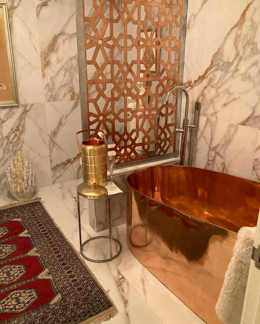 A luxurious bathroom featuring a Thompson Traders Baccarac copper bathtub, marble walls, a decorative screen, and a patterned rug.