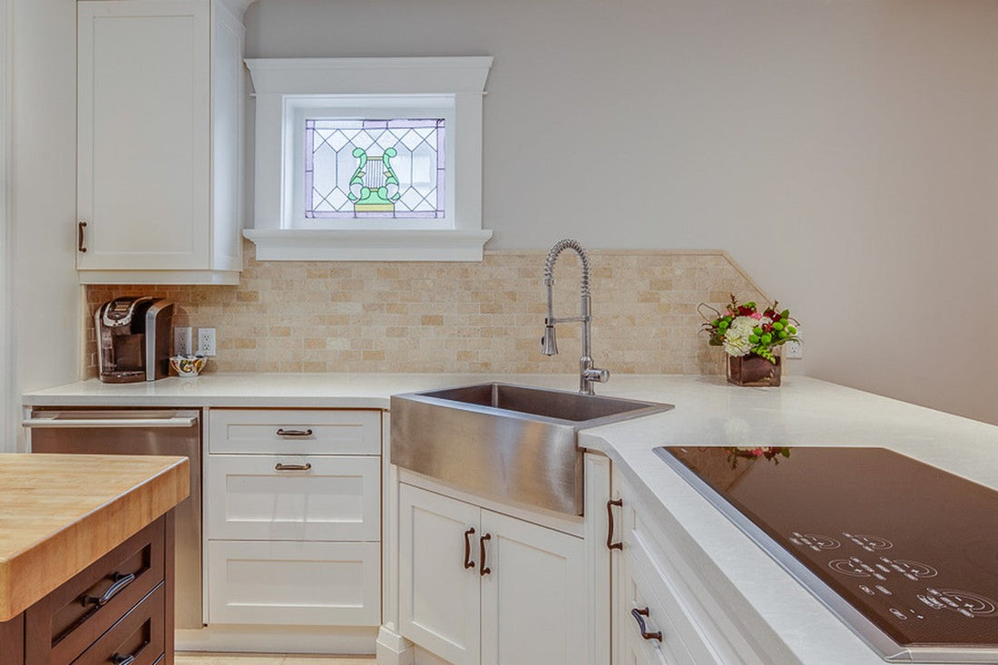 Stainless steel apron-front farmhouse sink in a white corner cabinet under a stained-glass lyre window