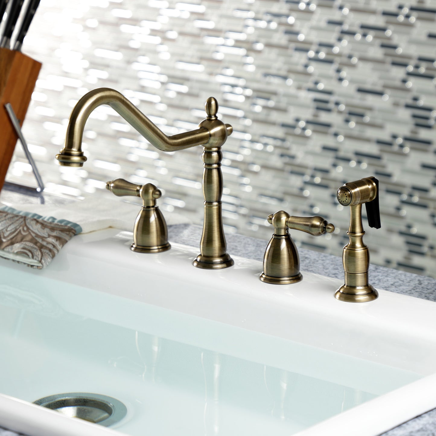 Kingston Brass "Antique Brass" widespread kitchen faucet set on a white sink with a decorative tile backsplash.