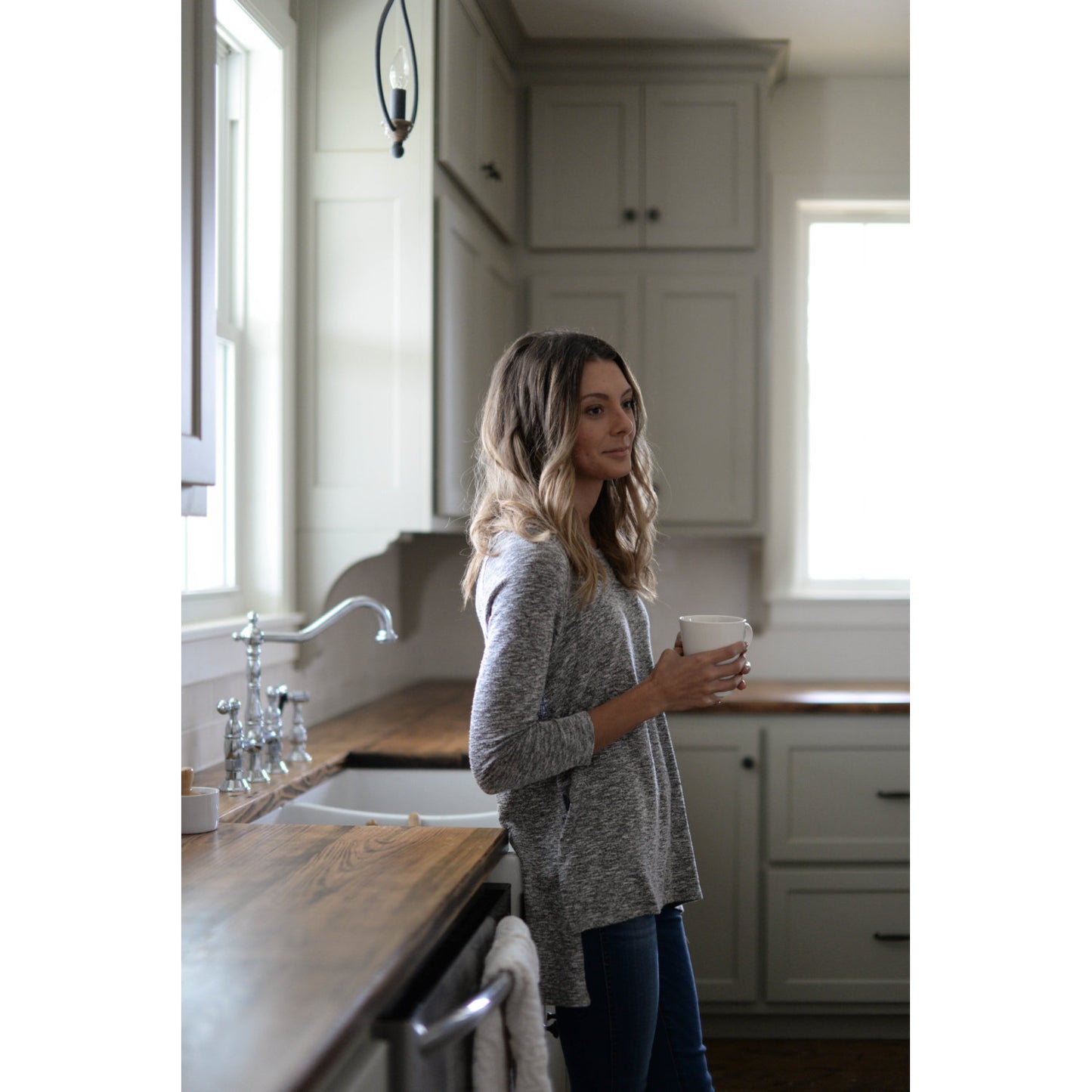 Woman enjoying coffee in a stylish kitchen featuring the KINGSTON Brass Heritage Bridge Kitchen Faucet with Brass Sprayer.