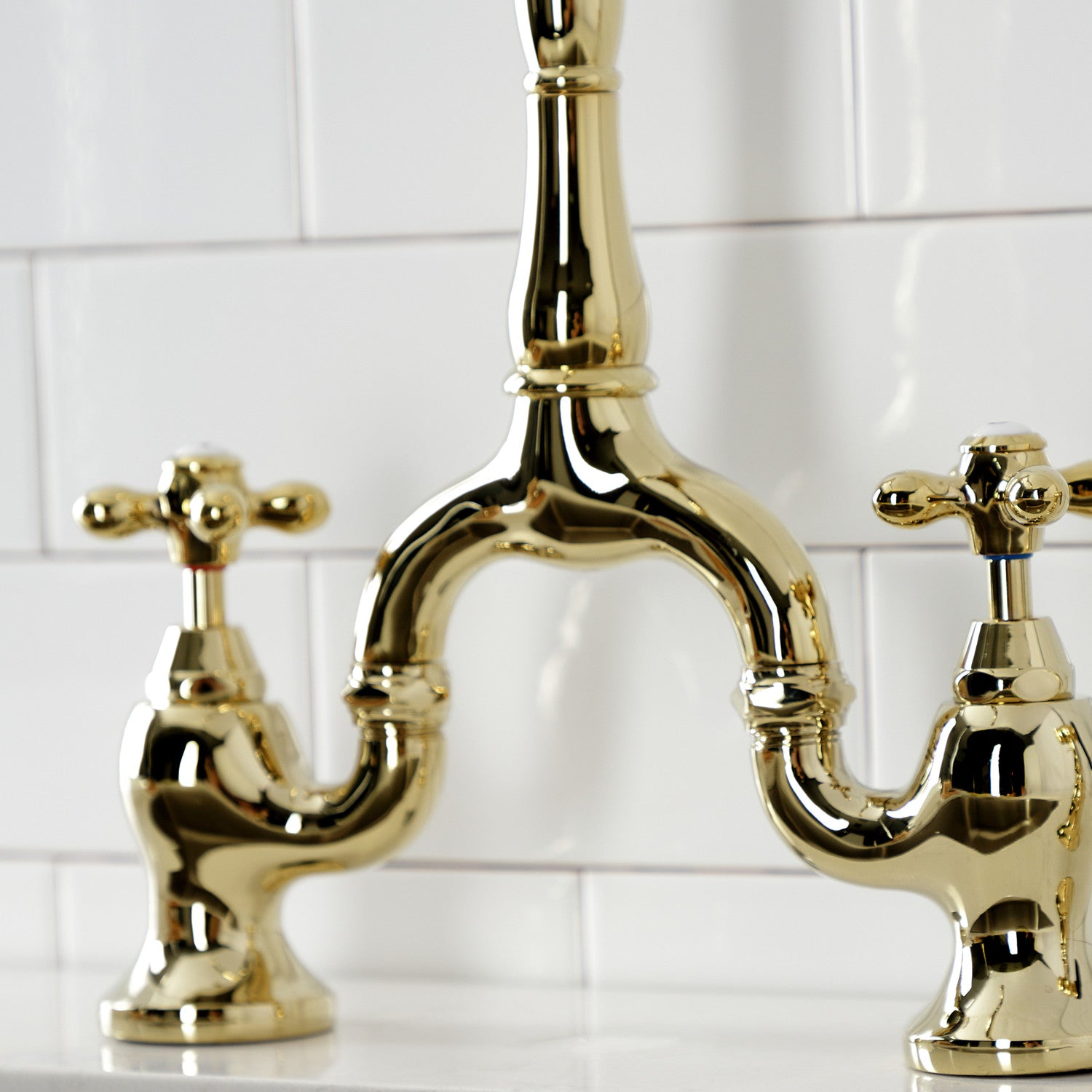 Kingston Brass KS7752AXBS polished brass bridge faucet set against a white tiled wall