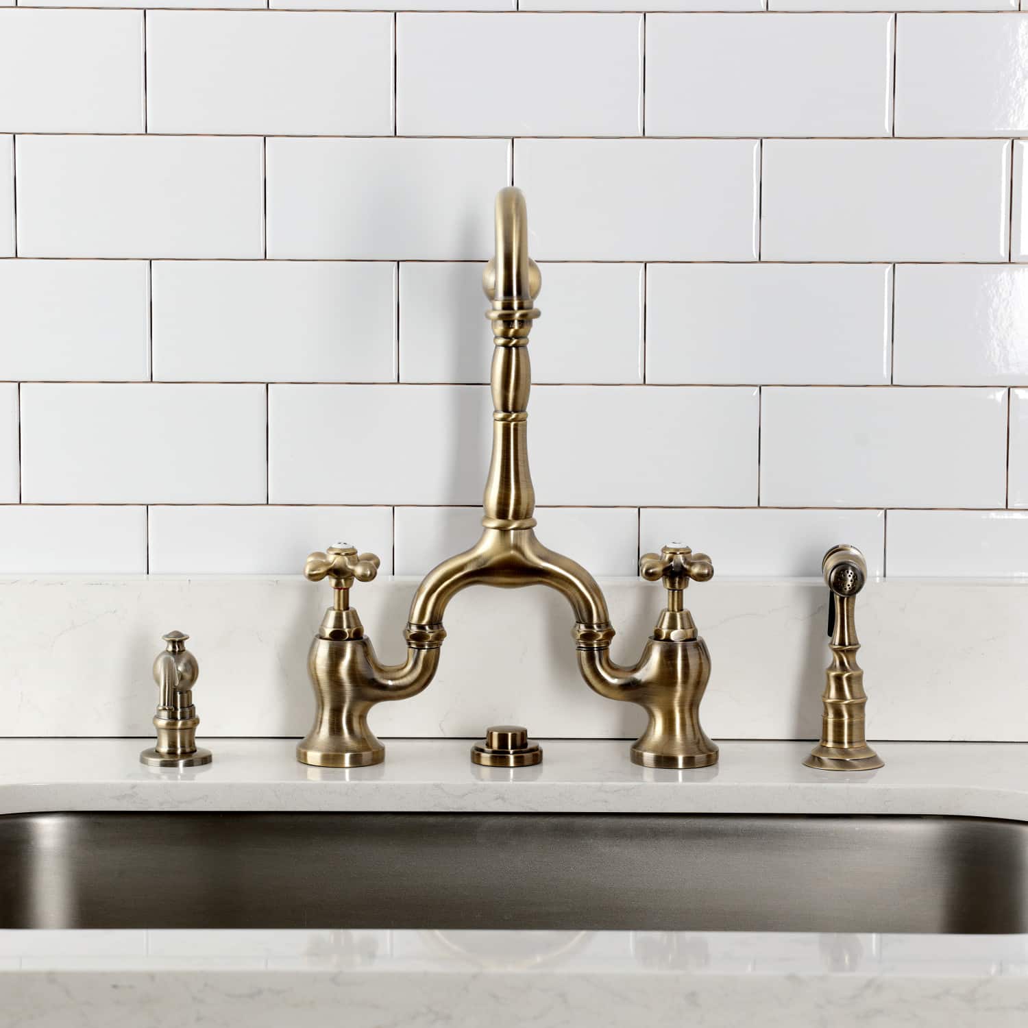 Kingston Brass KS7753AXBS Antique Brass  faucet installed over a kitchen sink with white tiled wall background