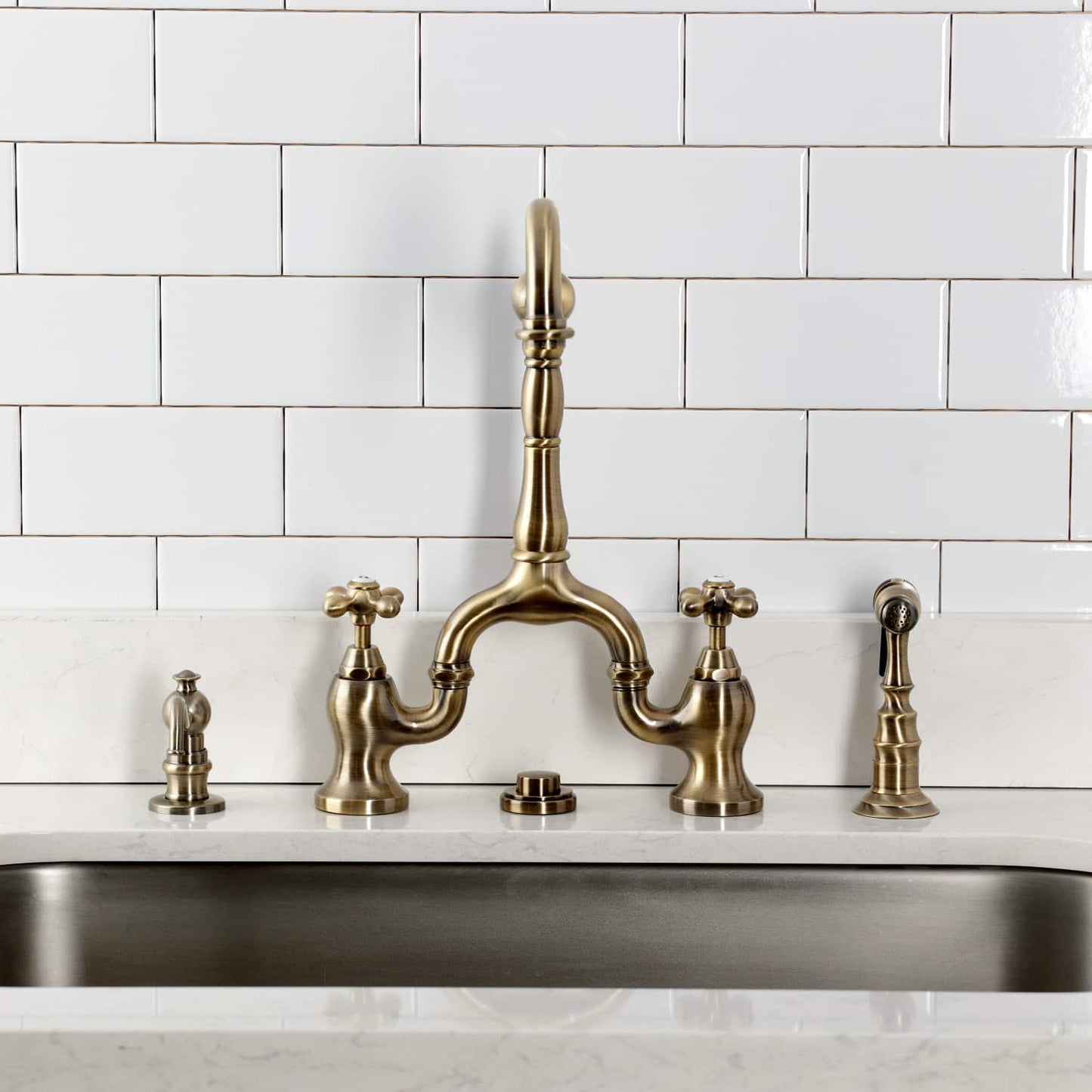 Kingston Brass KS7753AXBS Antique Brass  faucet installed over a kitchen sink with white tiled wall background