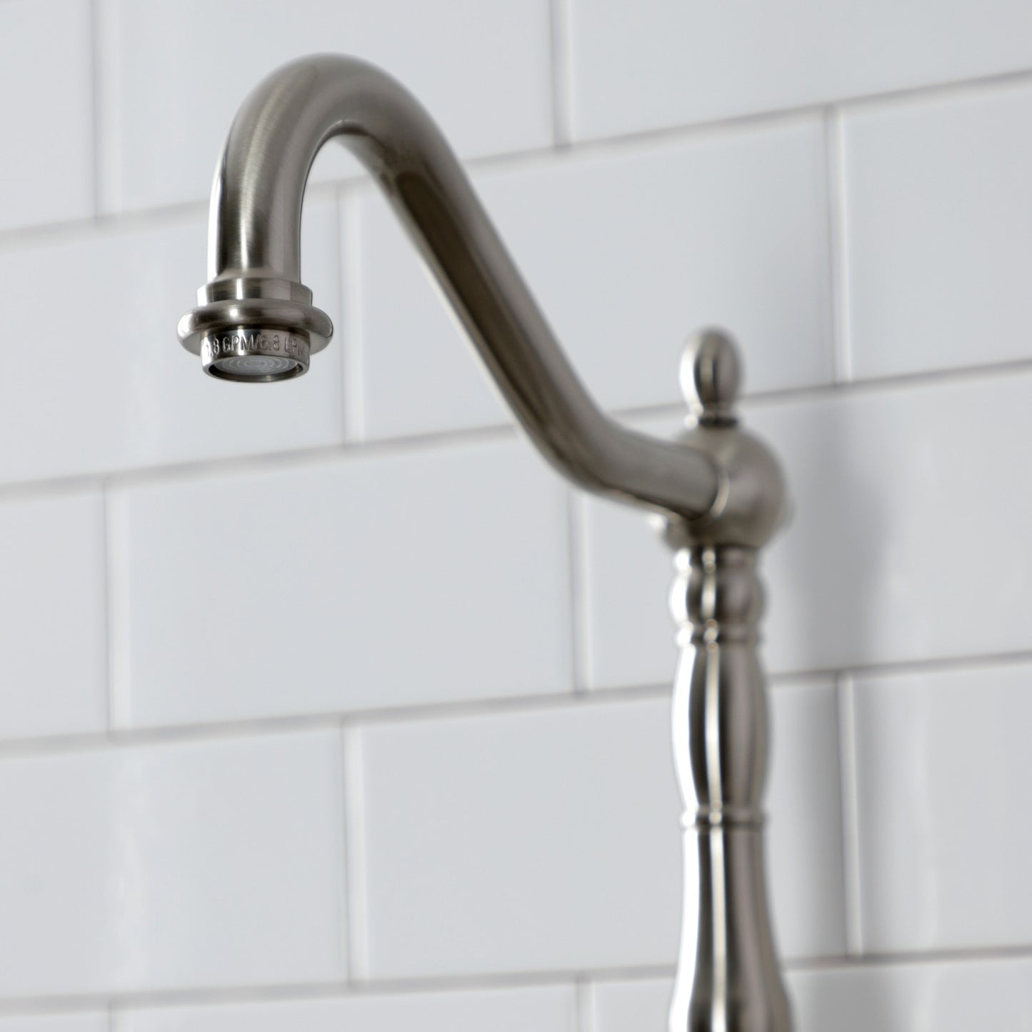 Close-up of the spout of Kingston Brass English Country Deck Mount Bridge Kitchen Faucet against a white tile background.