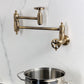 Kingston Brass KS3103RX Belknap Wall Mount Pot Filler Faucet, Antique Brass on a wall with a stainless steel pot below.