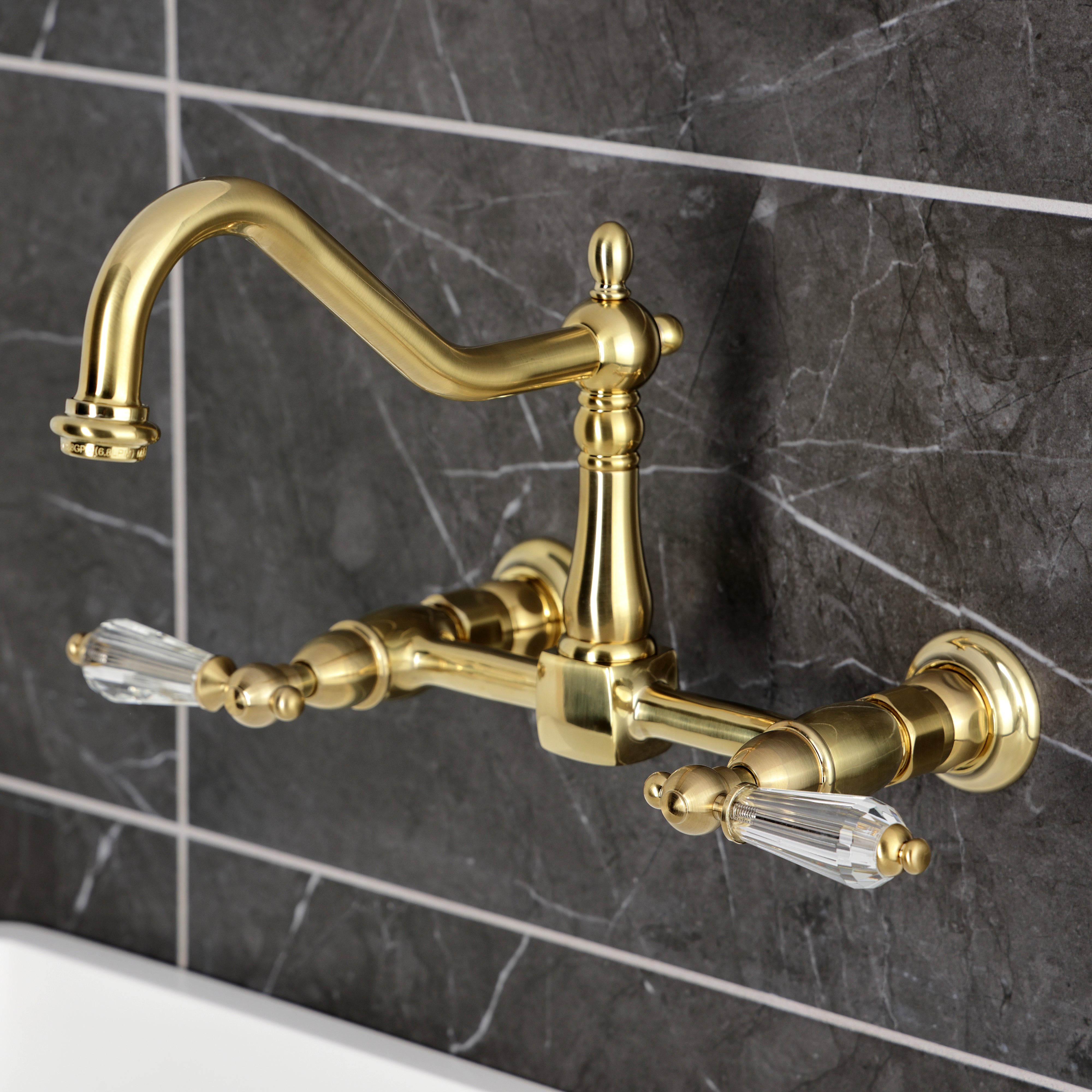 Kingston KS1243WLL Wilshire Two-Handle Wall Mount Bridge Kitchen Faucet, Antique Brass on dark marble background.