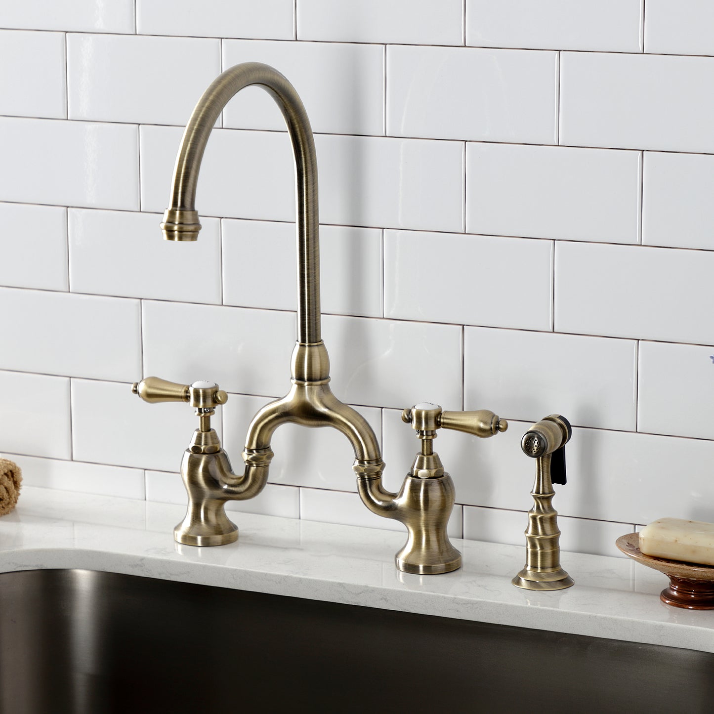 Kingston KS7793BALBS Heirloom Bridge Kitchen Faucet with Brass Sprayer, Antique Brass mounted on a white tiled kitchen wall.